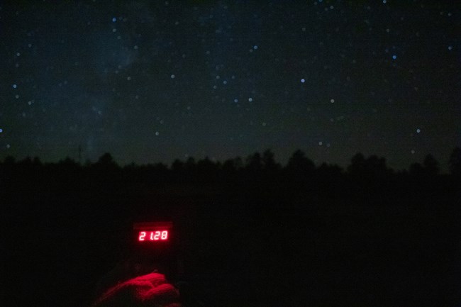 A device with the numbers 21.28 glowing in red is held up against a starry sky.
