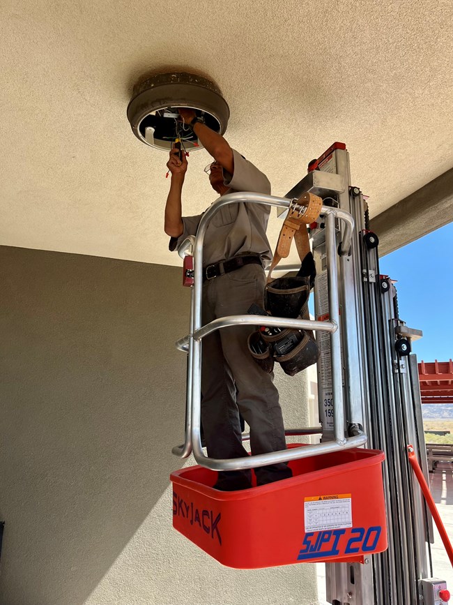 A maintenance person stands on a lift machine reaching up to work on wiring in a light fixture under a canopy of a building entrance.