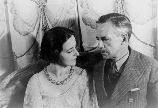 Portrait_of_Eugene_O'Neill_and_Carlotta_Monterey_O'Neill A black and white portrait of Carlotta Monterey and Eugene O'Neill. Carlotta, seated on the left, is wearing an elegant dress with a floral necklace. She has short, dark hair styled neatly and is looking intently at Eugene O'Neill.