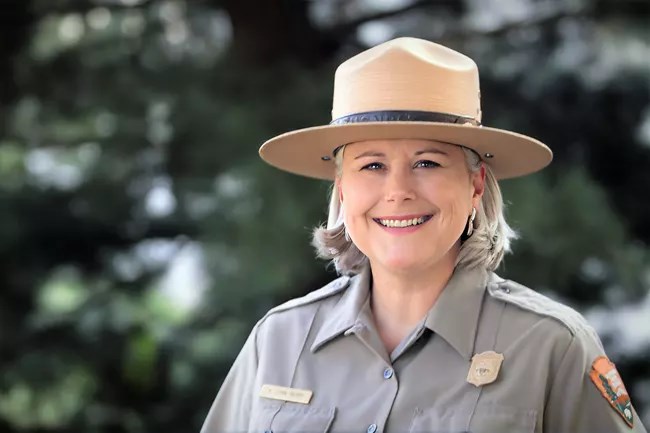 An older woman in a park ranger uniform is smiling at the camera. Trees in the background.