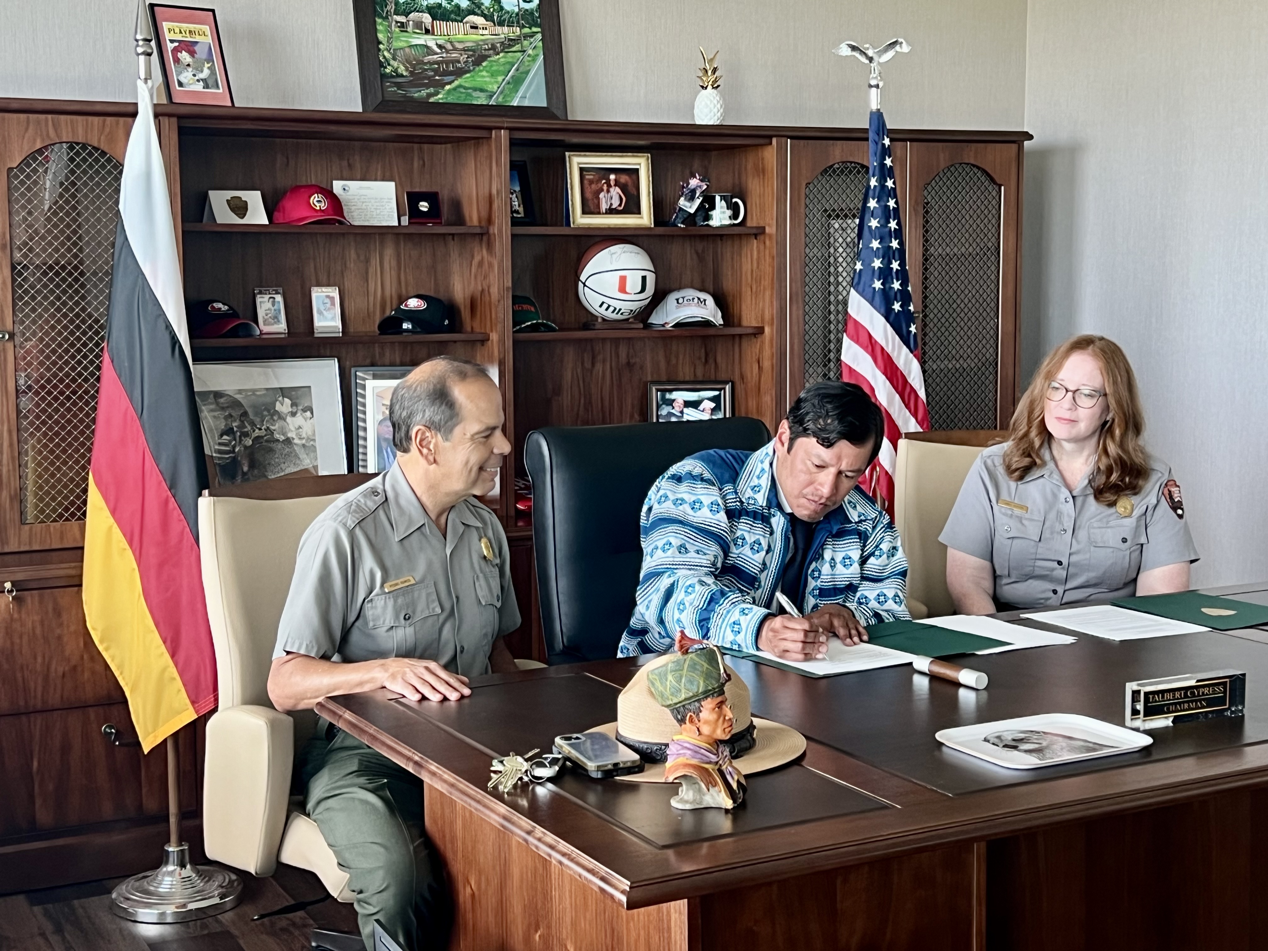 Three individuals sit at a desk flanked by the Miccosukee Tribe flag and the US flag. The individual in the center is signing a document.