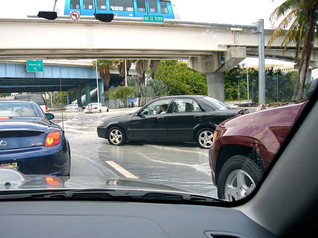 Looking out of the front of a car, the road is covered in inches of water. Several cars merge lanes. The street light is out. A bus travels over an overpass. In the background, below a green highway sign for the airport, a white car is in water up to the middle of the tires.