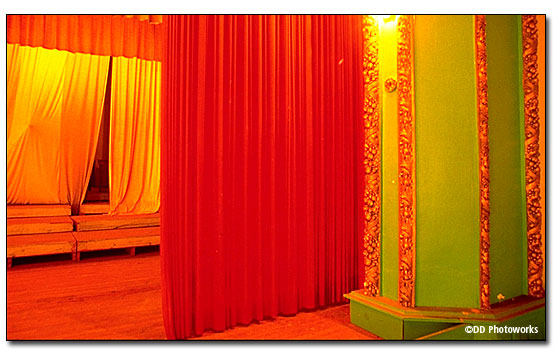 Color photo close-up of the stage details showing an octagon-shaped column painted bright green with gold leaf trim outlining each side of the octagon, with several layers of stage curtains behind. DD Photoworks