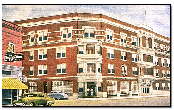 Tinted post card from around 1950. This is the same building that was first built by the Woodman of the Union, four stories tall, brick with light colored glazed terra cotta window surrounds and balconies. The building sits on the corner of two streets and the main doorway faces the corner. There are blinds on the street level windows, but no awnings at this time. To the left of the National Baptist building, across the street, is a pink colored brick building with a sign that reads 'Recreation Center.' There is a sedan parked in front of that building and one on the side street by the National Baptist. There are two men standing in the doorway of the National Baptist.