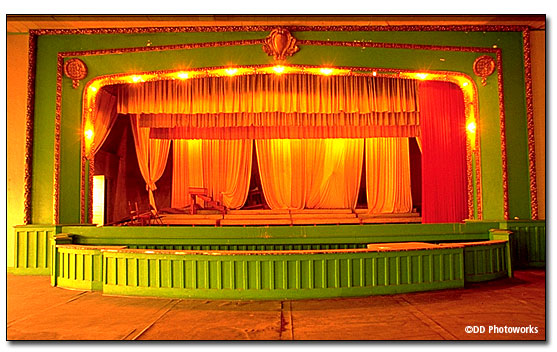 Colored photo showing the full frontal view of stage including the orchestra pit. Above the stage curtain there is a large round medallion at either end and centered above the stage is a cartouche, all covered with gold leaf. The red velvet curtains are open, stage lights are on and there is a part of a three-tiered riser on stage right. DD Photoworks