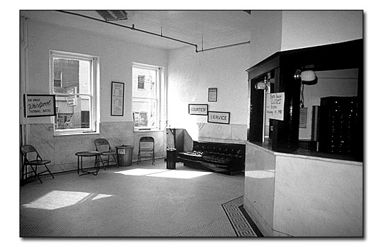 Black and white photo with the hotel lobby office in an enclosed booth on the right. There are two bare windows looking out to the Malvern Ave. side of the building, several folding chairs against the wall next to the windows, an upholstered bench sits against the wall to the right. You can see some detail of the mosaic tile border around the office booth-it has a classical Greek design.