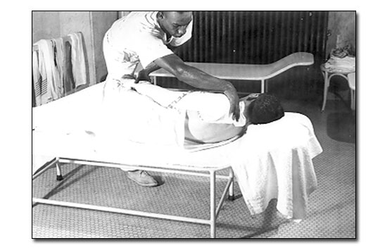 Black and white photo showing two pack 'tables.' The pack table is more like a padded cot with the head end raised slightly. In the foreground is a white man lying on his side on the pack cot. He has a white bath sheet wrapped around him and the African American pack attendant is putting a towel on his shoulders. There are towels hanging on a bar on the wall on the right side. The pack attendant is wearing a white t-shirt, white pants, and tennis shoes. On the back wall you can see a radiator and on the back right is a small armless chair with towels stacked on it.