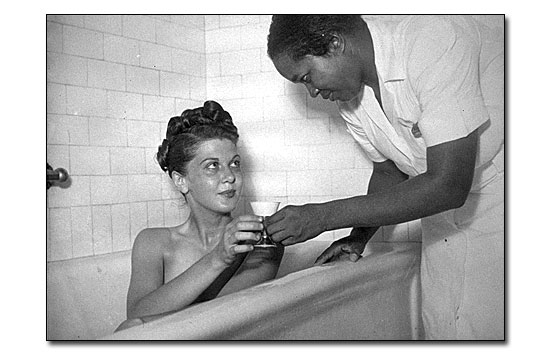 Black and white photo showing a close up of a white woman in a deep bath tub. An African American bath attendant is handing her a cup of water from the right. The attendant is wearing a white uniform dress similar to a nursing uniform of the time.