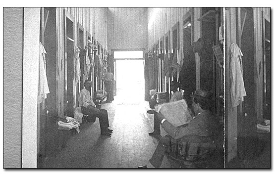 Black and white photo looking from one end of the room to the other, with a wide aisle down the center and bath stalls on each side. There are men sitting on benches outside the stalls with towels stacked next to them. In the right foreground sits a man in a suit with hat reading a newspaper; he is thought to be the photographer.