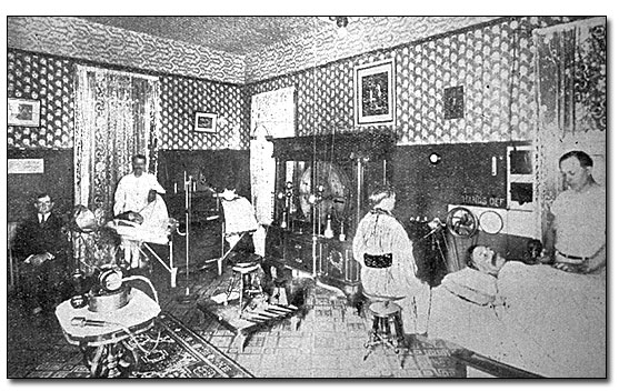 Black and white photo of the men's massage department in the Palace Bathhouse around 1900. The large room with dark wainscoting two-thirds up the wall with large patterned wall paper above and on the ceiling has several stations with various electro-therapy equipment and massage table. Counterclockwise from the right foreground there is a white man with a mustache on a massage table, covered by a sheet, with the masseur standing behind the table. The masseur is a young white man dressed in a white t-shirt and light colored pants. Next there is a man with a flowing silky robe sitting in front of a piece of electro-therapy equipment. Then there is a large glass enclosed cabinet on the wall with several things on shelves, possibly batteries. On the back wall,  there is a curtain hanging that may be covering a doorway. Next there is a man wearing a white lab coat at a small table and sitting next to the table is a gentleman fully dressed with a coat and tie. In the left front there is an electrotherapy device on a stand.