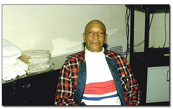 Mr. Payton is an African American  man, sitting casually in a chair in front of a credenza. He is wearing a white turtle neck shirt with a red and blue checked quilted shirt over it. Behind him on the left end of the credenza is a stack of blankets and some books covered in plastic. To the right of the credenza you can see part of TV cart.