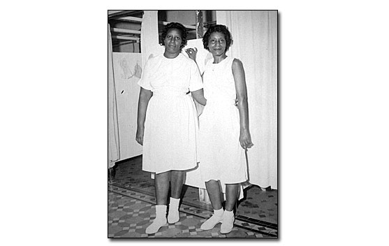 Black and white photo of two African American female bath attendants.  They are standing in front of dressing stalls that have curtains across the doorway. The stall partitions are white as are the curtains. The women are wearing a white short-sleeved uniform dresses that comes below the knees, white anklet socks and white tennis shoes.