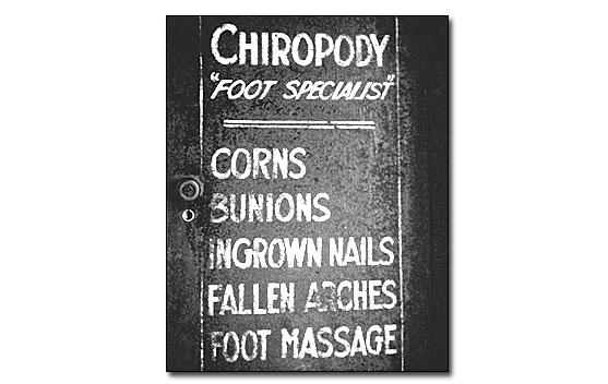 This sign is painted on the outside of the electrical fuse box outside the Chiropody Room. The box cover is black and the lettering is gold leaf. In all caps it reads, in a vertical row, Chiropody/'Foot Specialist'/Corns/Bunions/Ingrown Nails/Fallen Arches/Foot Massage.