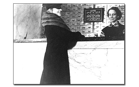 This black and white photo shows a nicely dressed white woman standing at the Fordyce Bathhouse counter with a clerk behind the counter. The woman is wearing a winter coat with a fur stole collar, hat, and gloves. The clerk is a younger white woman. The counter is tall, coming up higher than the waist and made of marble. Behind the counter on the wall is a bank of brass lock boxes and there is a sign with the bathhouse rates hanging on the lockers.