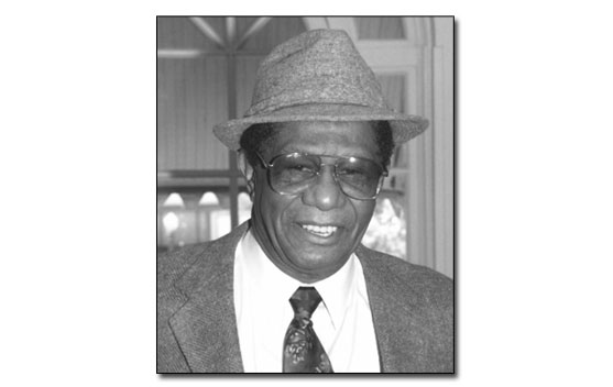 Black and white head and shoulders shot of a smiling Mr. Puckett. He's an African American man wearing a tweed jacket, white dress shirt and tie and a tweed fedora.