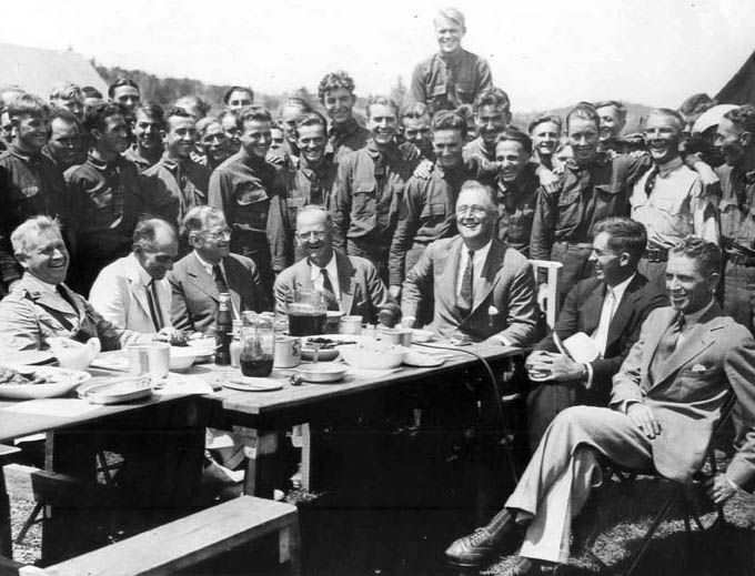 FDR at a table with the CCC boys in the background.