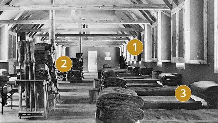 Beds lined against the wall of a barracks building with a stove in the center of theh room.