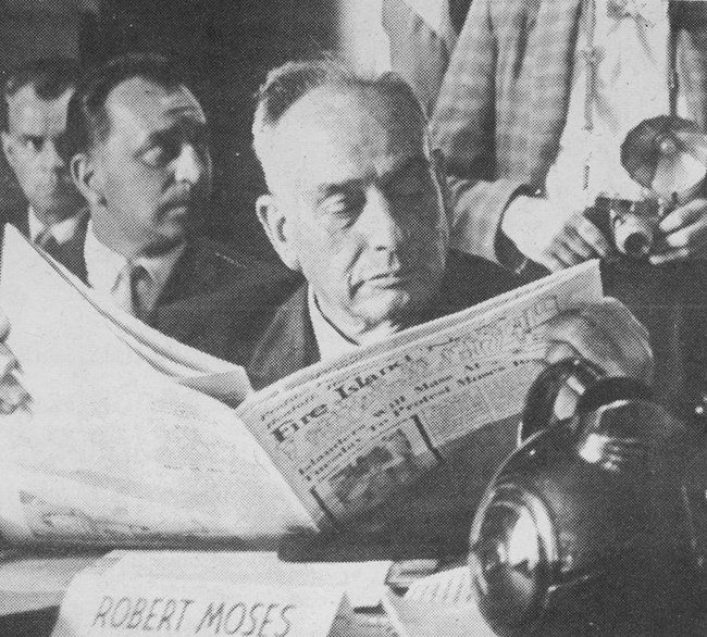 Robert Moses sits at a dais and reads a newspaper.