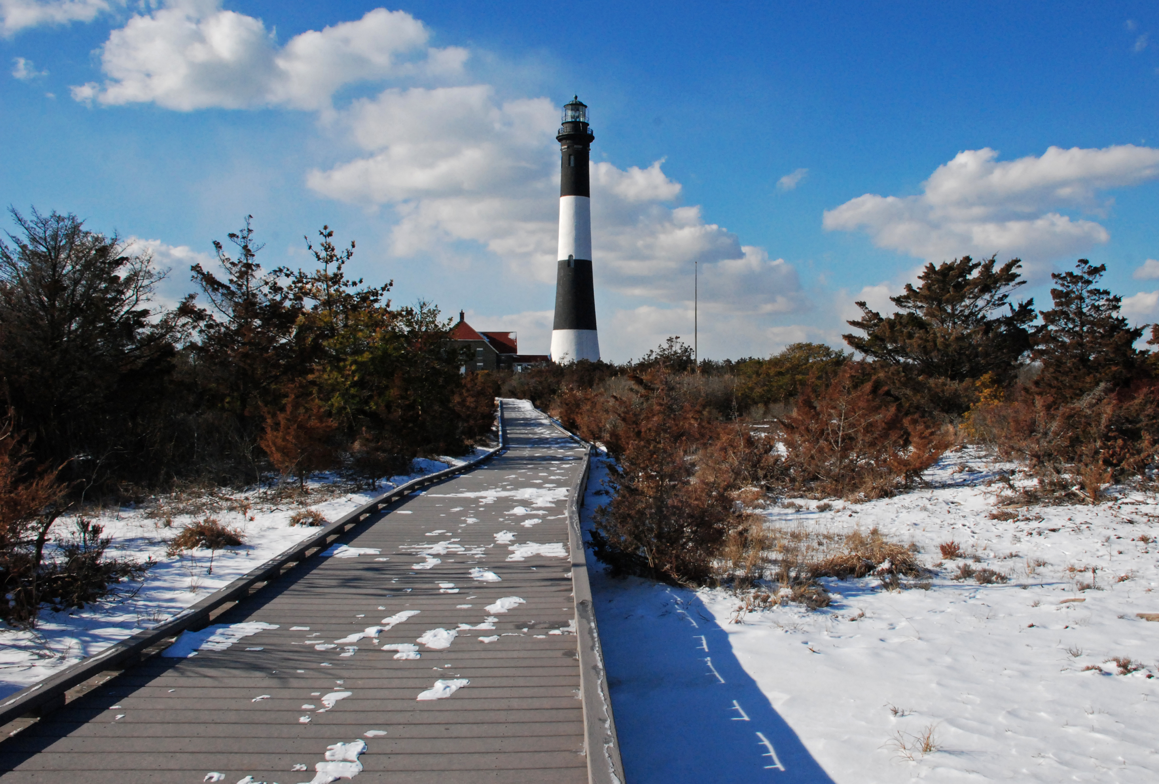 A snowy boardwalk leads to a black and white lighthouse tower.