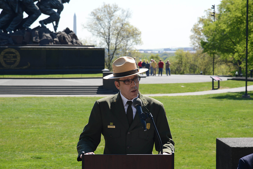 A man wearing a green National Park Service Uniform with a wide-brimmed hat, leans over a podium in the center of a grassy field between a couple trees.