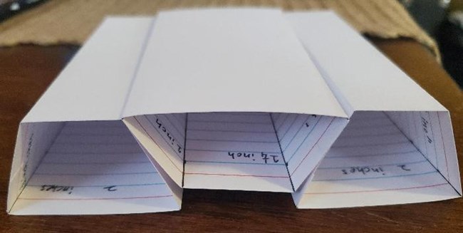 Index cards have been made into four sided blocks. The outer two are sitting on their broadest sides and are holding up the third broad side up. Tops are all at the same level.