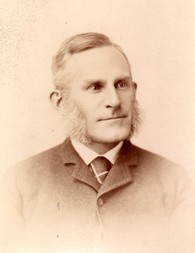 A historic photograph of a man with large mutton chops looking right.