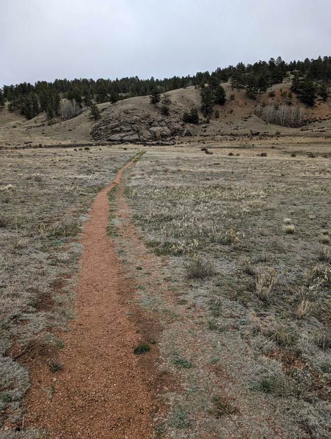 A reddish gravel trail heads toward a rocky hillside covered in tan grass and green trees. The sky is overcast with gray clouds.