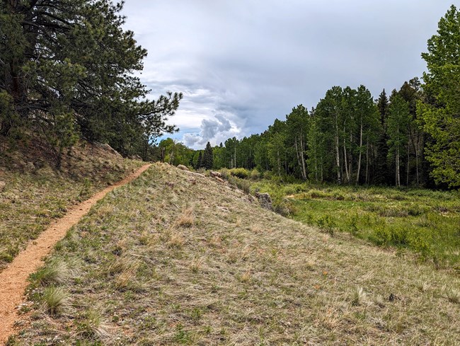 The Twin Rocks trail follows a gently rising grass covered hill. Below the hill sits a verdant riparian area. In the distance, storm clouds swirl around a single point of blue sky.