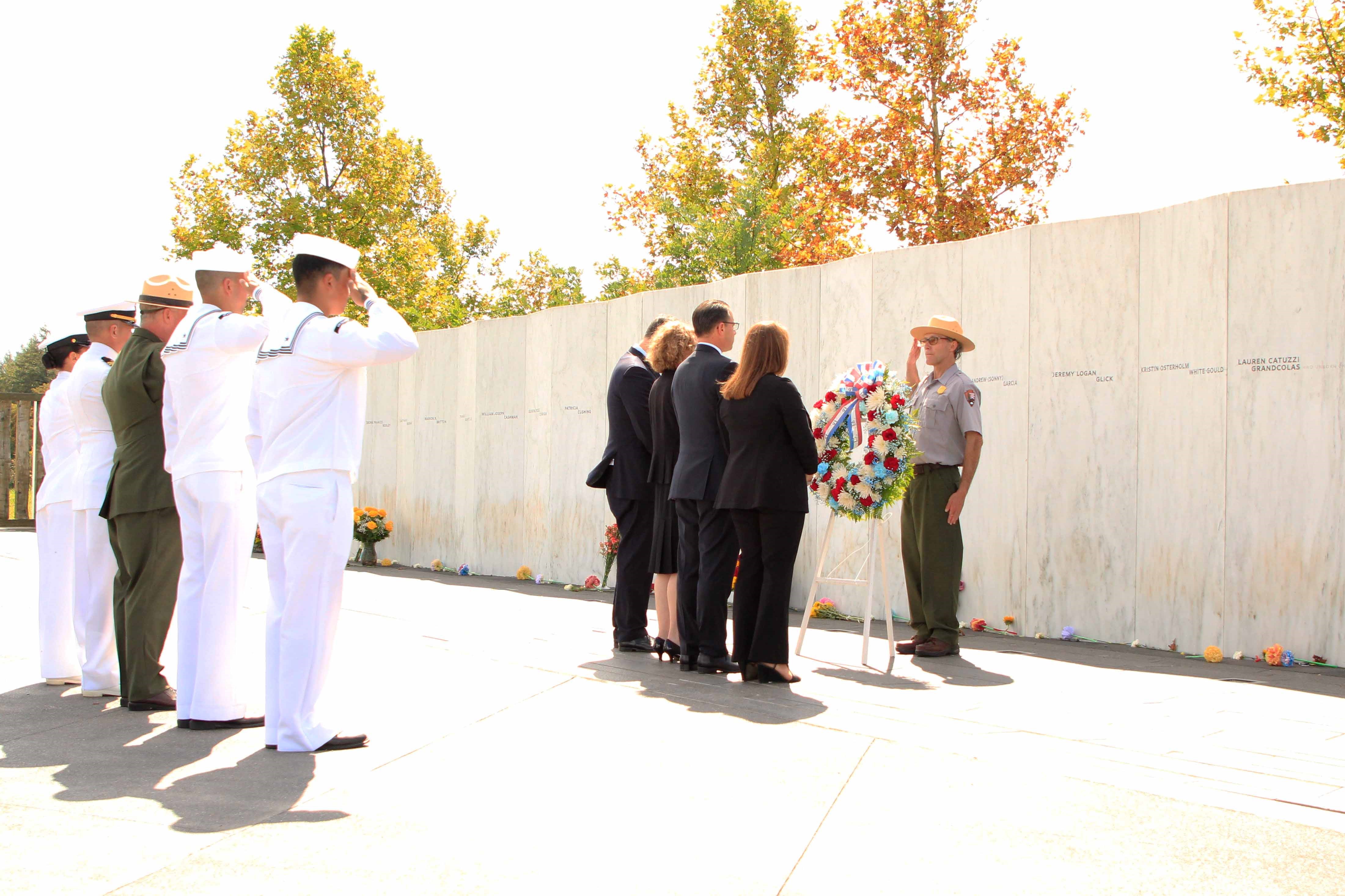 A park ranger in uniform salutes and stands in front of a wreath.