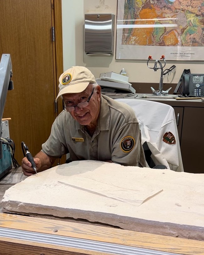 A man in a tan uniform holds a pencil-like tool to a large rock and smiles.