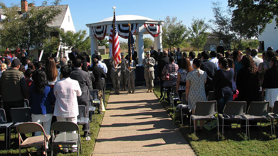 View toward stage at naturalization ceremony. New citizens on the side, color guard in the center, well canopy and trees in the background.