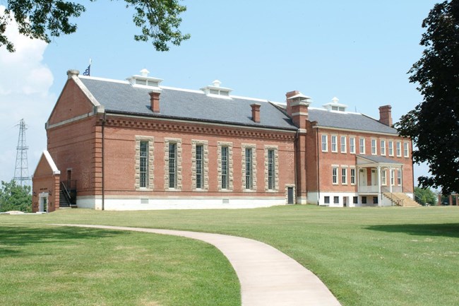 The red brick visitor center building with long, barred windows with white stone on all sides. A paved path leads to the entrance through green grass.