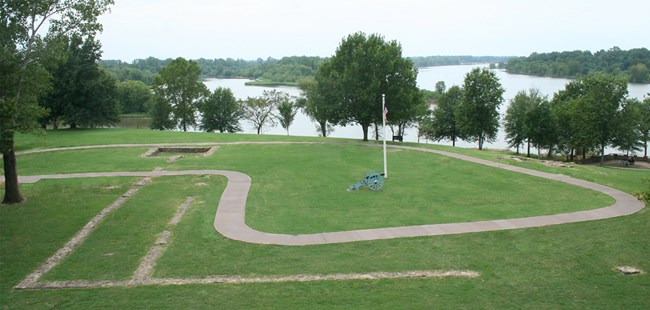 A sidewalk winds through gray stone lines in grass with trees and a river in the background.
