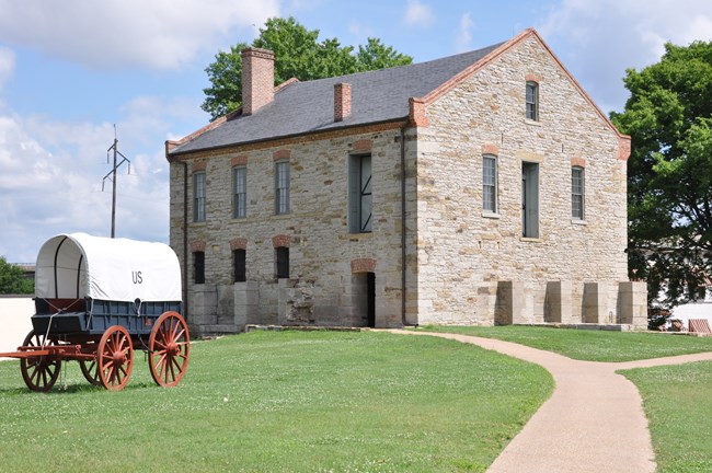 A three-story gray stone commissary building with a oaved walkway leading to its door and a white and red canvas supply wagon in front.
