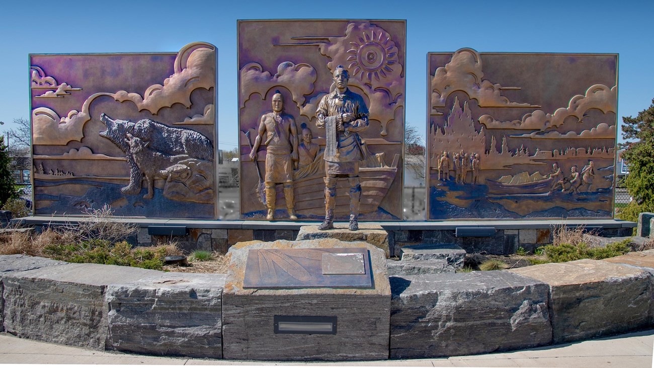 3D5BD9EC-C1E6-83C1-5C350FBC5C9D906F 3 shiny brass panels with animals and natives in relief. In front, an Oneida man stands with feathers adorning his head and a wampum belt in hand.