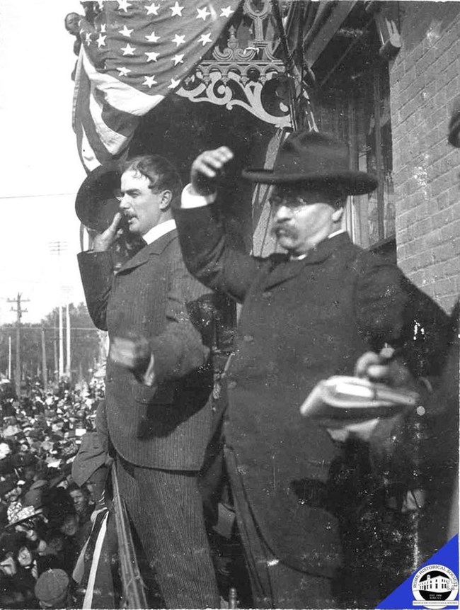 A side view of a man with a monocle in a nice suit, waving to the crowd with his hat.