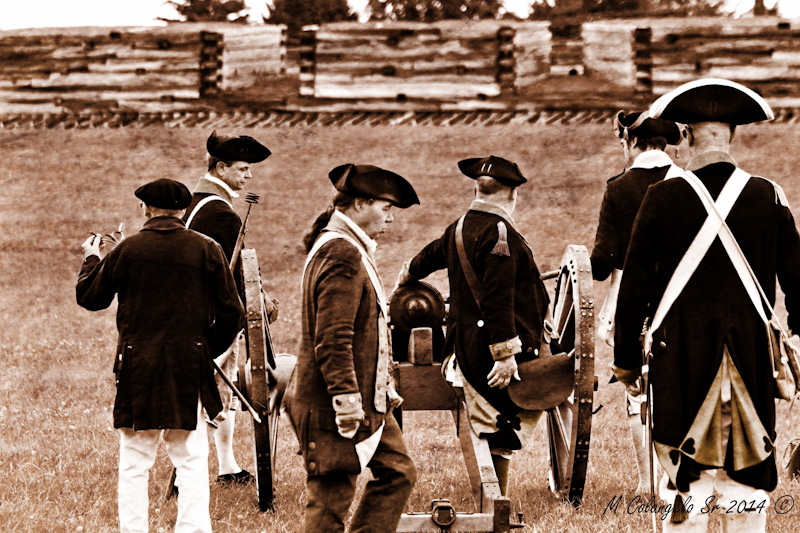 In a photo, men in British-style uniforms stand around a cannon facing the fort wall. 