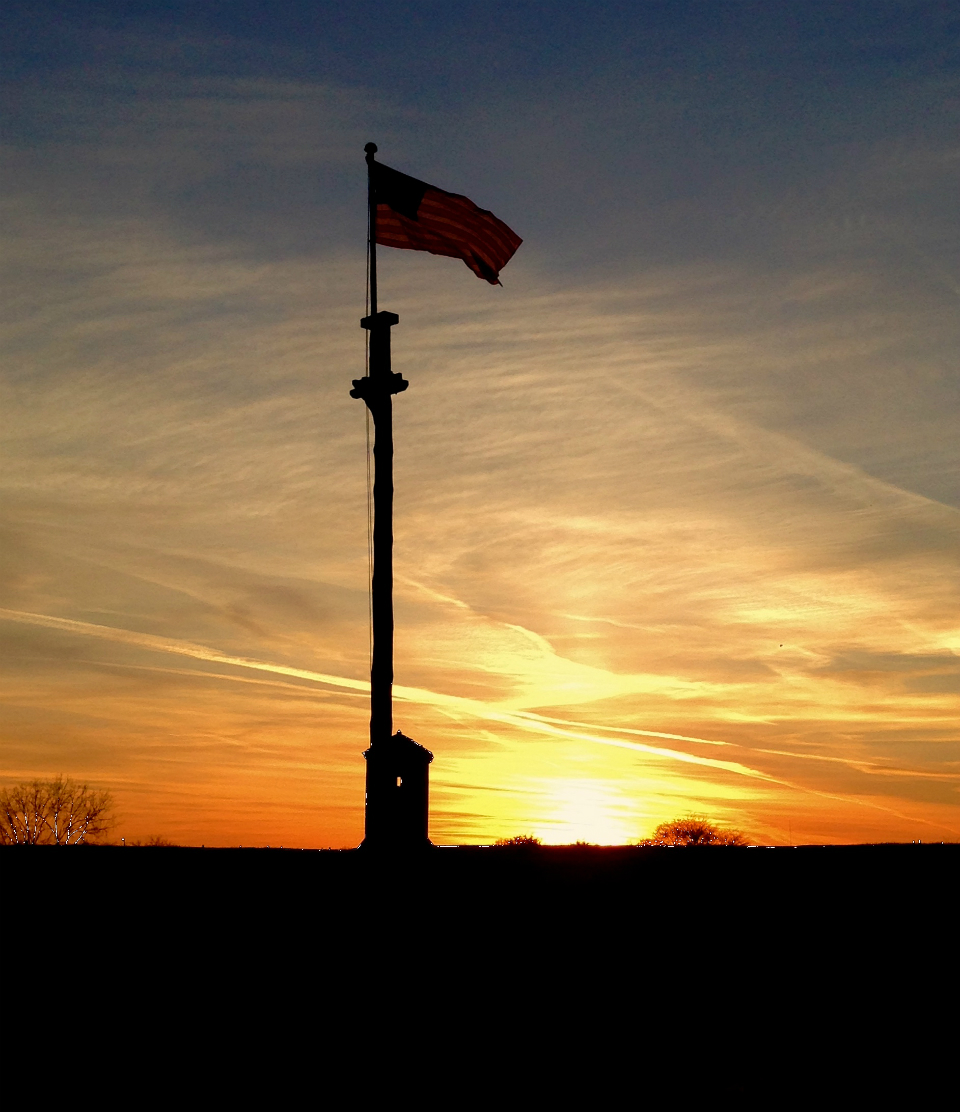 A flag waves in a vivid orange sunset over a shadowed wooden wall.