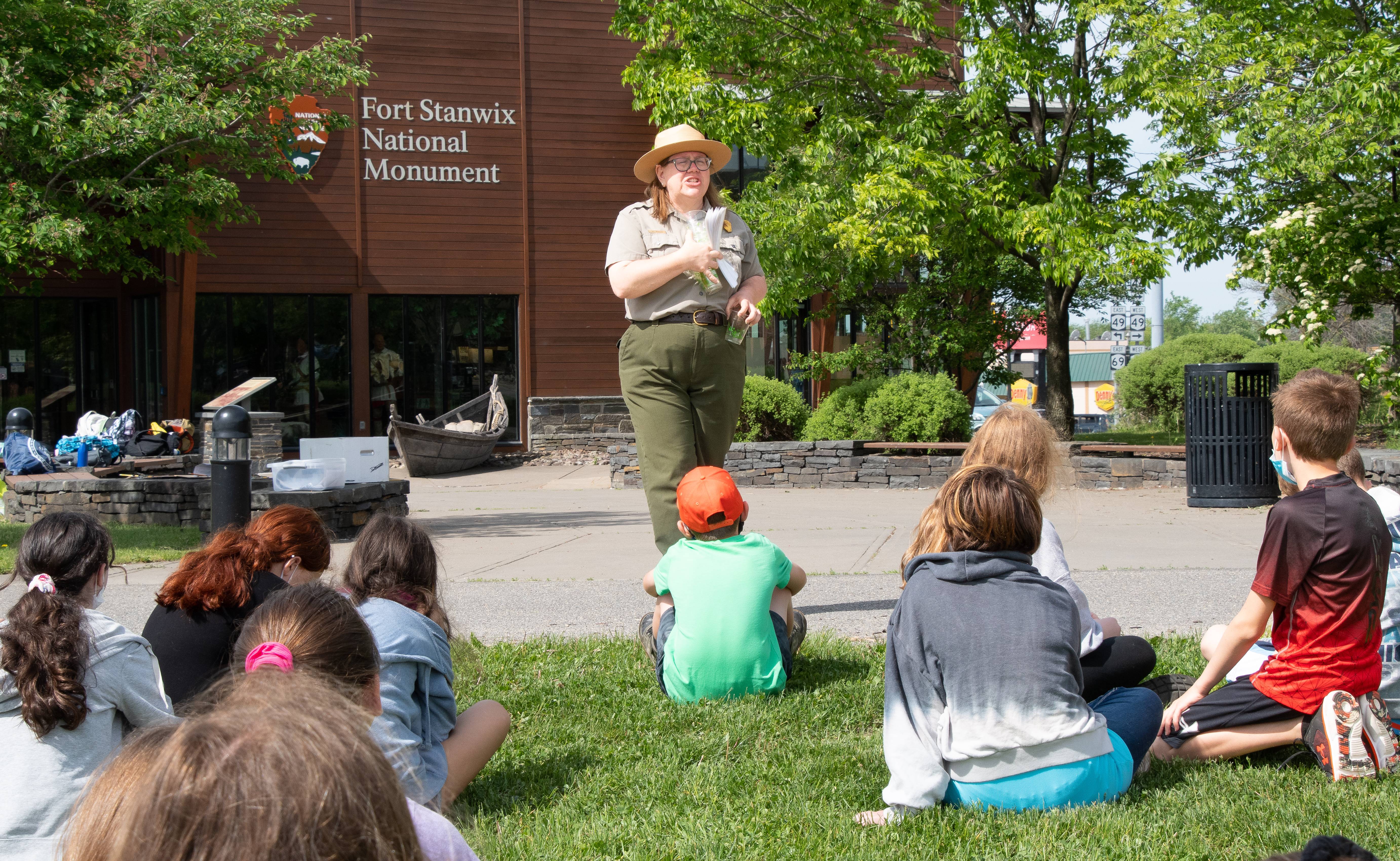 A park ranger stands in front of a group of children seated on the ground in a very verdant area. You are looking at the ranger over their heads.