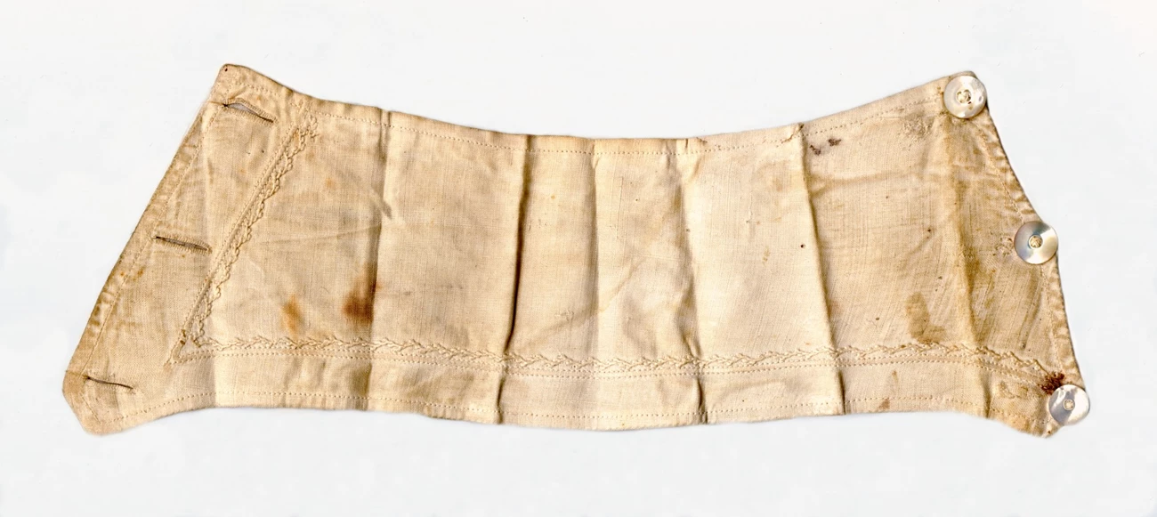 Laura Keene's Bloodstained Cuff Off white sleeve cuff with an embroidered pattern, three buttonholes on one side, and three pearl buttons on the opposite side, with folding creases and dark red-brown stains