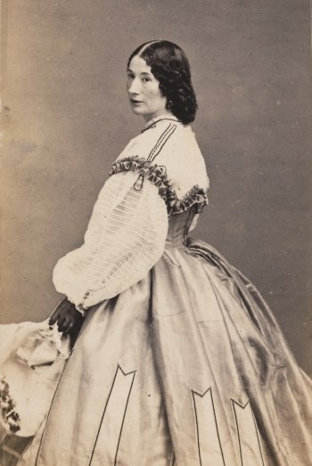 Black and white photograph of a white woman with dark curly hair standing in an elegant hooped dress.