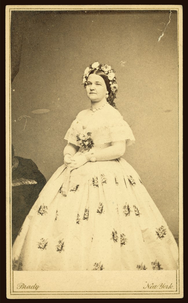 Sepia tone photo of Mrs. Lincoln wearing white gloves, a floral wreath, billowing light-colored dress with a repeating floral design and lace blouse with flowers on the front..