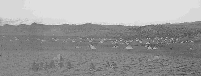 A few people sitting in the foreground with tipis in the background in front of mountains