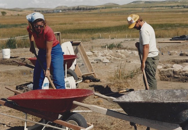 A woman and a man working the archaeological digs with shovels and wheelbarrows. A Woman, in red shirt, blue jeans, sunglasses and headscarf. A man in a baseball hat, white shirt and green dungarees.