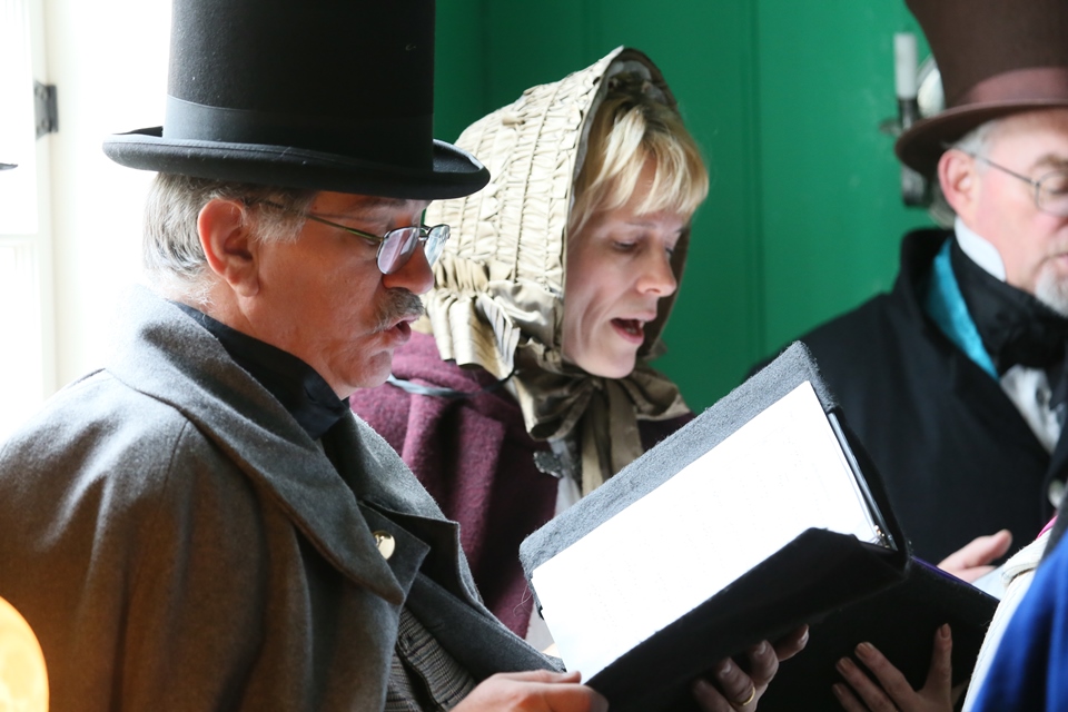 Three carolers in period clothing sing Christmas carols from a book.