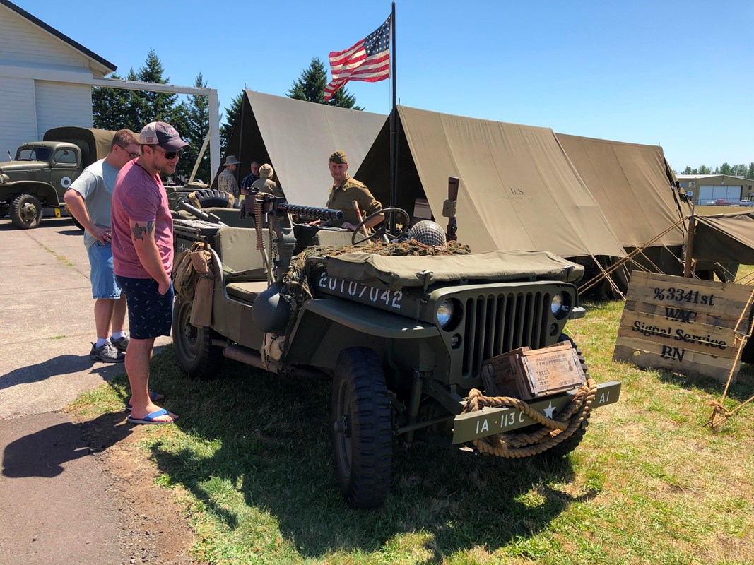 Two men look at a WWII Jeep in front of army tents and an American flag while a man in WWII army uniform looks on.