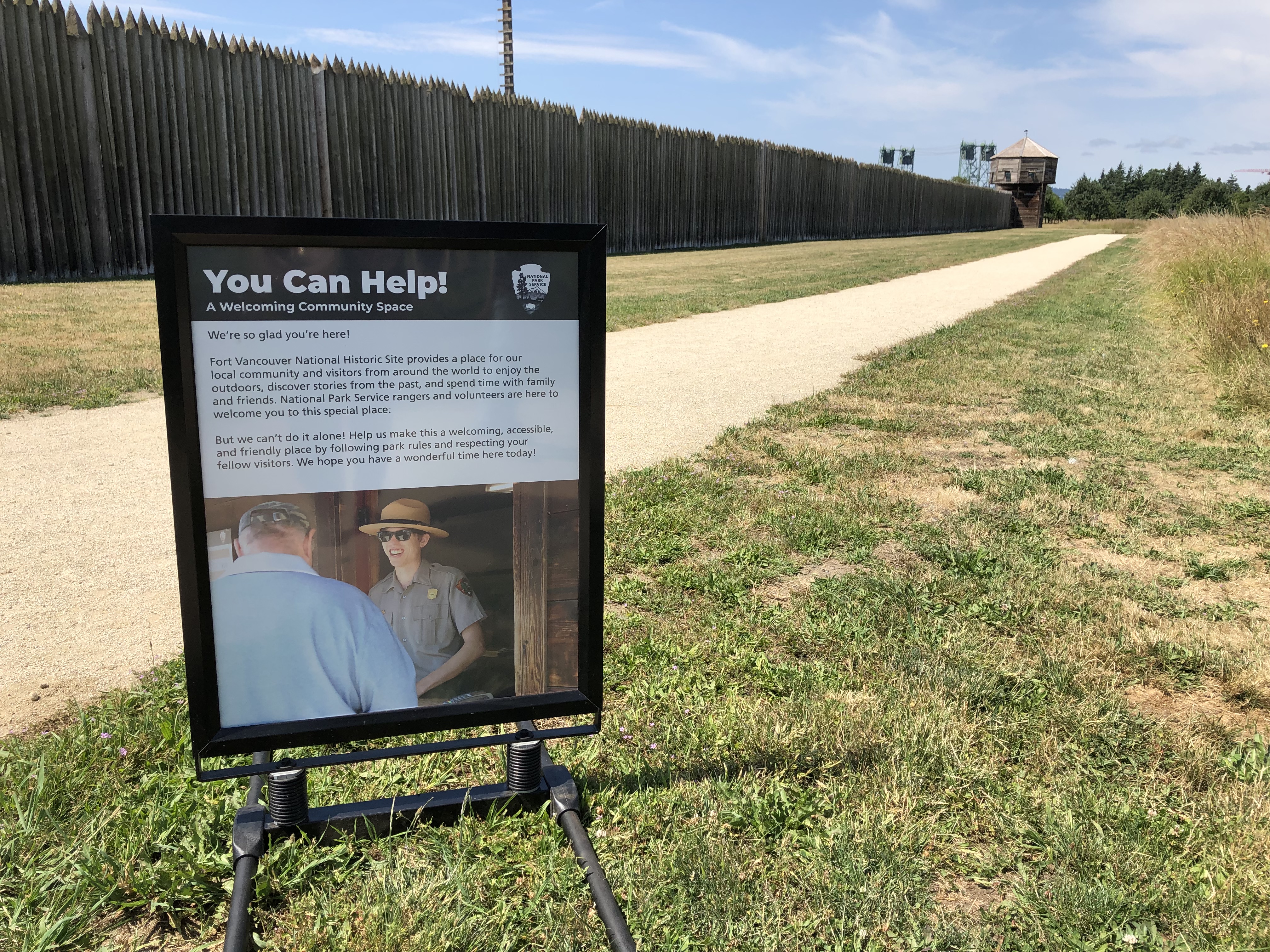 A panel with the headline text "You Can Help" sits in a grassy area outside Fort Vancouver.