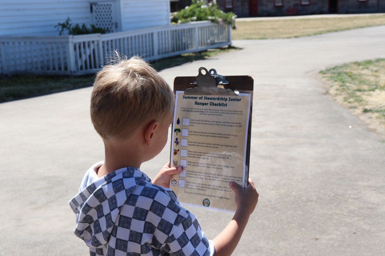 A young child holds up a clipboard with a Summer Stewardship Junior Ranger Checklist inside Fort Vancouver on a sunny summer day.