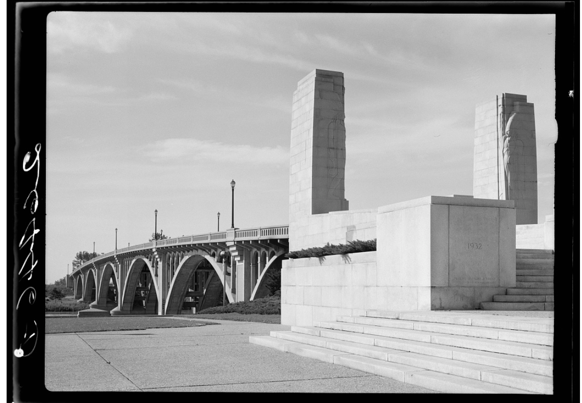 A black and white photo of the bridge approach, a set of light gray granite stairs and two gray granite columns with 1930's stylized Native American figures carved on them