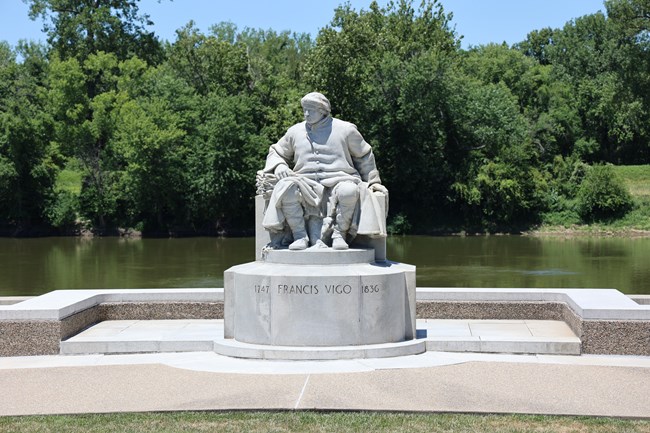 A gray granite statue of a man wearing a long coat and moccasins sitting on a bench. The statue sits on front of a river.